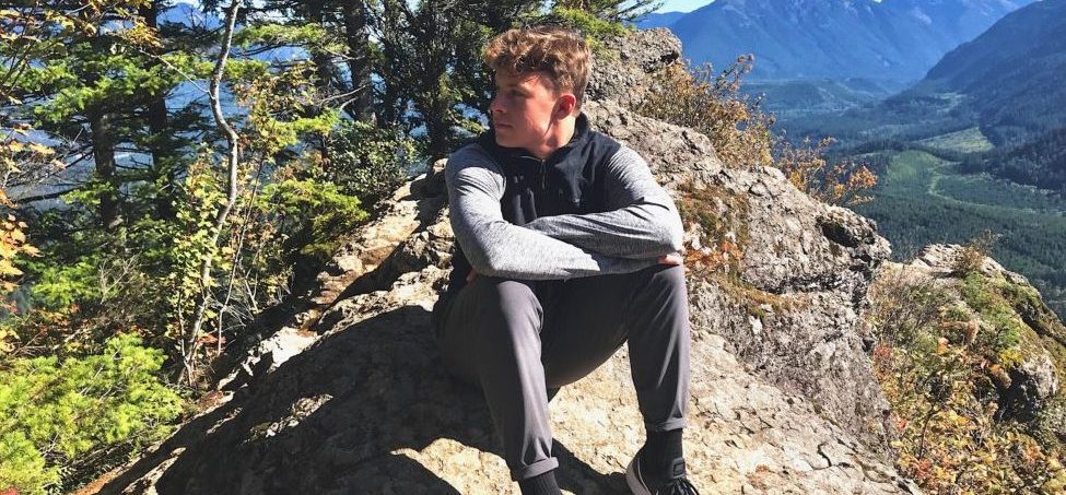Michael sits on a rock on a hiking trail with mountain views behind him.