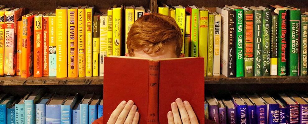 A person holds a book to their face with color coded books on a shelf behind them.