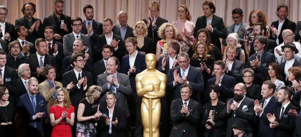 A person-sized oscar stands in front of a group of mostly white actors and film people