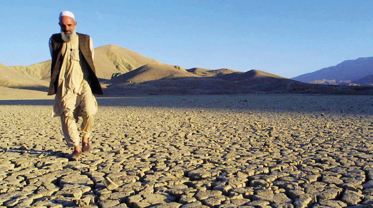 A man walks along dry, cracked earth with a mountain rising behind