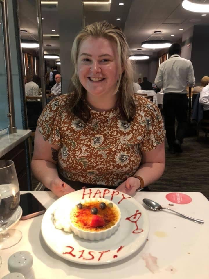 Lauren smiles, seated at a table in front of a crème brûlée. Around the plate, the words "HAPPY 21ST!" are piped in chocolate alongside a depiction of a martini glass