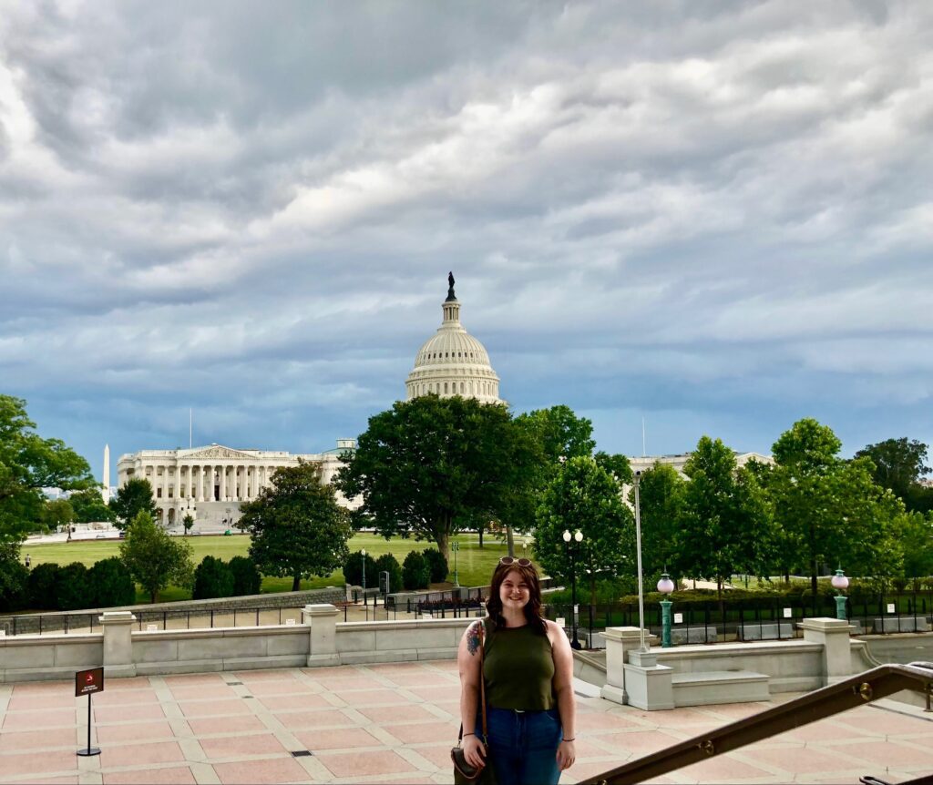Lauren stands in front of the United States Capitol building on a brick tiled courtyard. The building is partially blocked by green trees