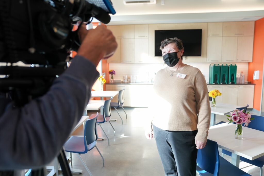 Jen Muzia stands in the Ballard Food Bank looking at a TV news camera.