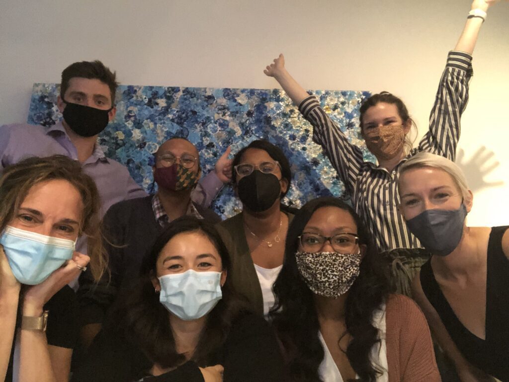 The Minerva team smiles in face masks with LaKita from CoCreate Work. The team stands in front of a multicolored blue background, and Kayla waves both hands in the air