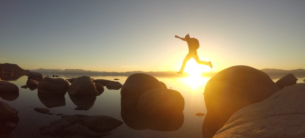 A person leaps from one boulder to another over water, with the setting sun in the background.