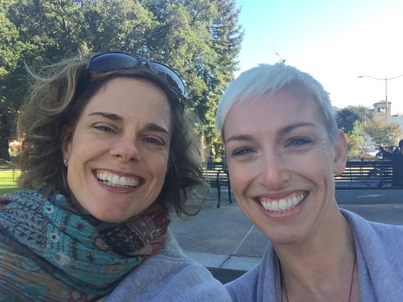Joy and Sara smile. A tree and bench are visible in the background.