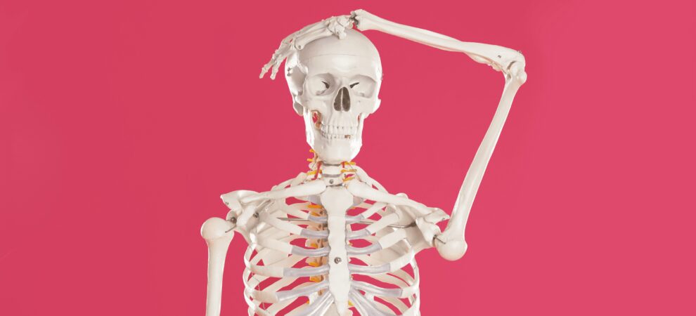 An image of a skeleton with one hand on its head in confusion against a pink background.
