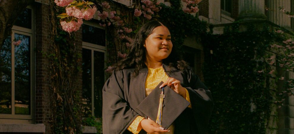 Falisha stands in the sun wearing a graduation gown and holding her cap. She is smiling off to the side.