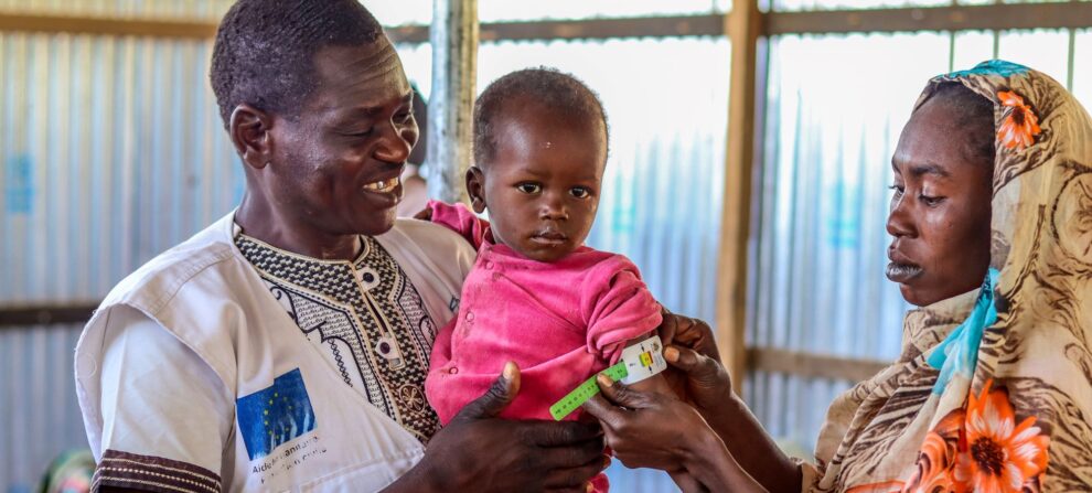Health worker, who is from Chad, holds a child while the child's mother measures their arm circumference with a tool called a MUAC band. Health worker is wearing a white vest, the child is wearing pink pants and shirt, and their mother is wearing a beautiful, printed wrap.