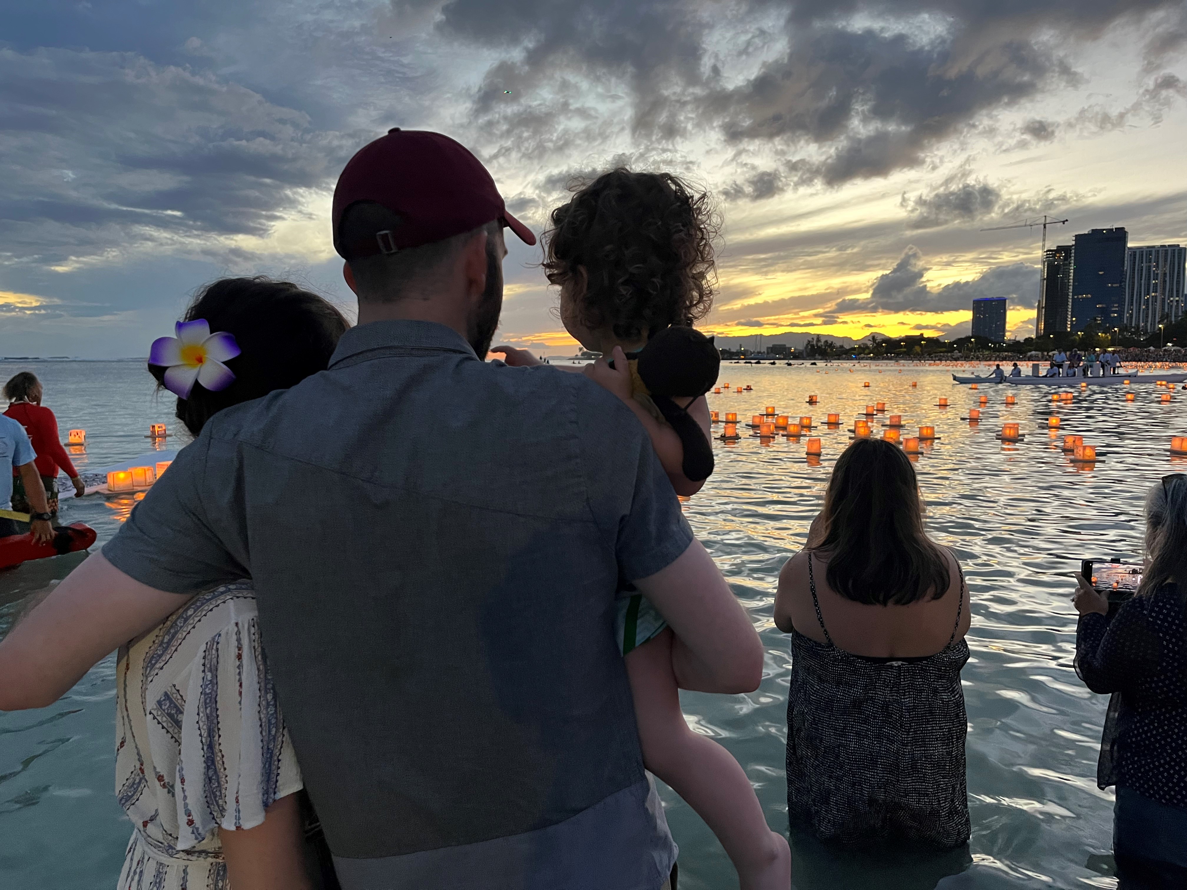 Kayla's family stands at the water watching the lanterns. Her husband is holding her daughter and they all have their arms around each other