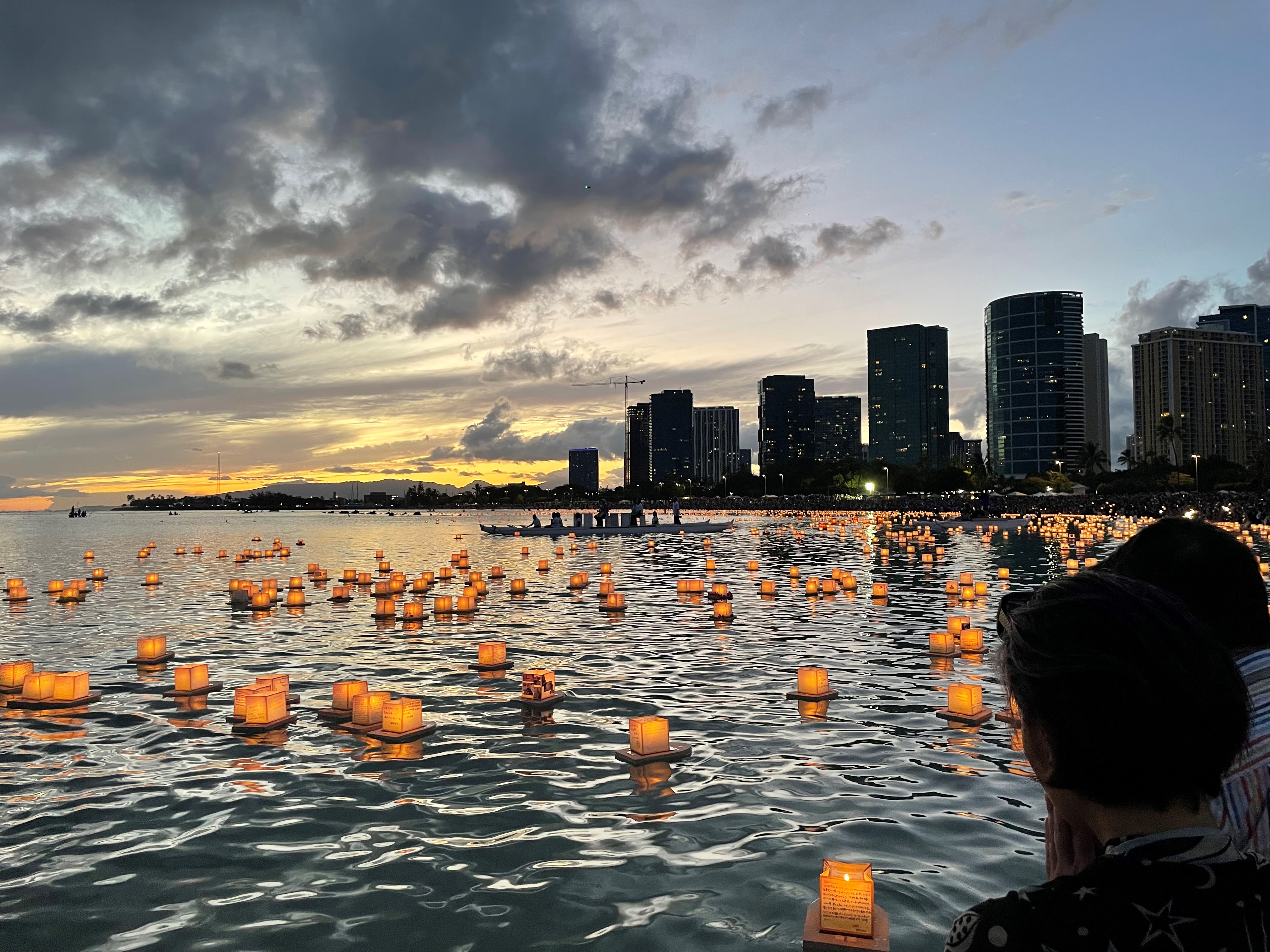 Photo of illuminated lanterns floating on the ocean with the cityscape of Honolulu in the background. It is night and the sky is dark with some sunset visible.