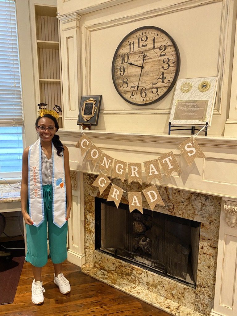 Elise stands by a banner that reads "Congrats Grad" wearing a stole that says "Minerva MVP"