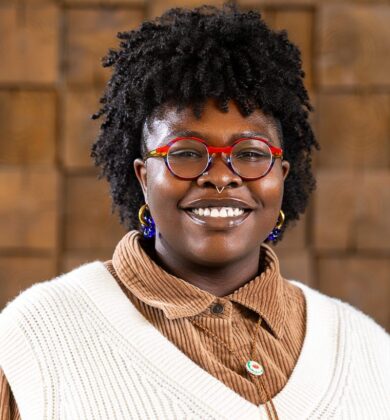 marissa is a dark-skinned Black Woman with kinky hair that is shaved on the sides. She has nose piercings and is wearing round, multicolored glasses. She also has on a sweater vest layered over a brown corduroy button down, with a bolo.