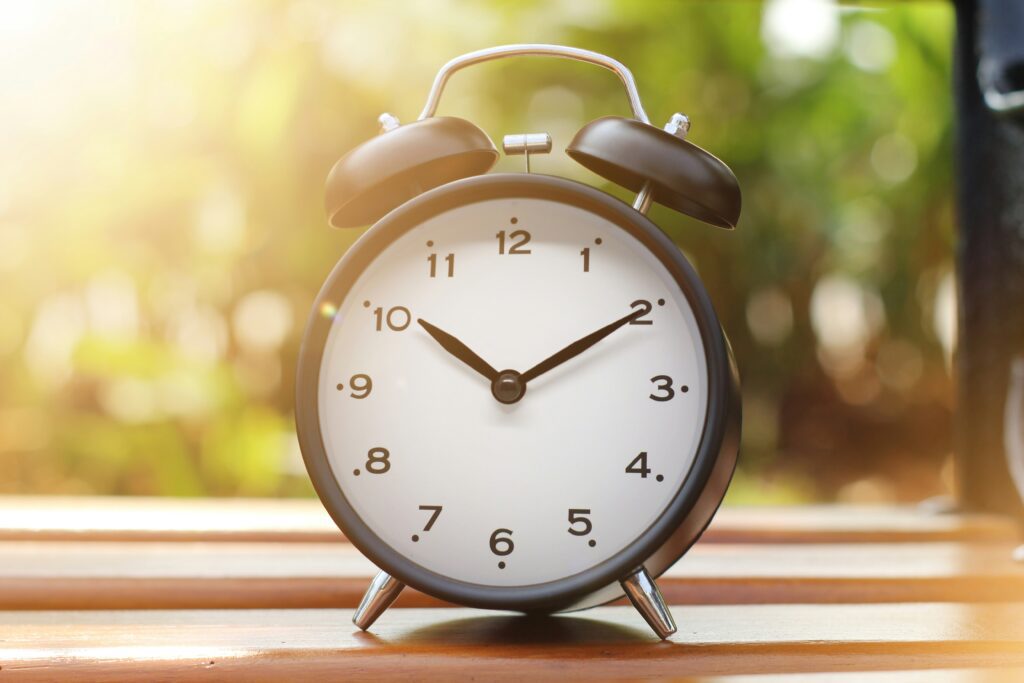 An old fashioned alarm clock sits on a window ledge. Outside the window shows a sunny day in soft focus.