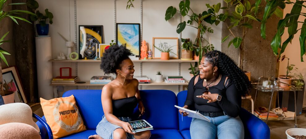 Morgan and Tobi sit on a blue couch, smiling. Morgan holds an iPad on her lap, and Tobi holds a notebook in mid air.