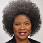 Tanya has a medium-dark skin tone and smiles in front of a beige background.