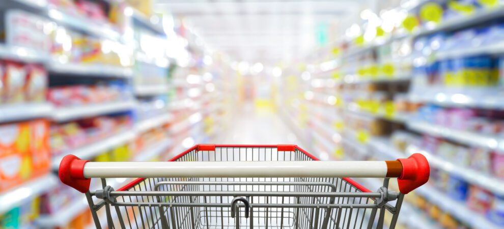 The view from a shopping cart entering a brightly lit aisle of a supermarket. Colorful products line the shelves.